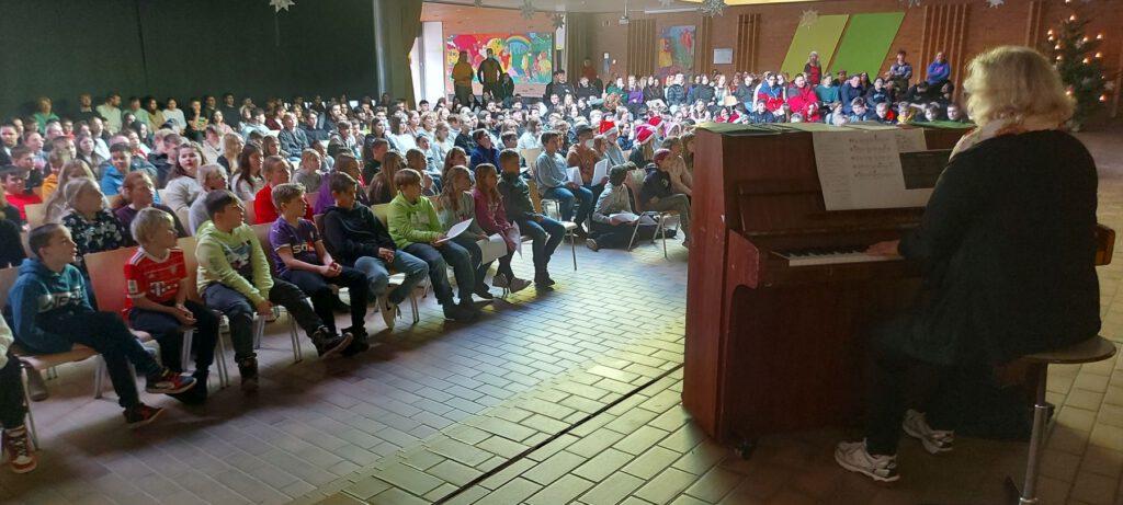 Frau Petermann sitzt am Klavier und spielt die Weihnachtslieder für die Schulgemeinschaft, die auf Stühlen in der Aula sitzt. Alle singen gemeinsam Weihnachtslieder.